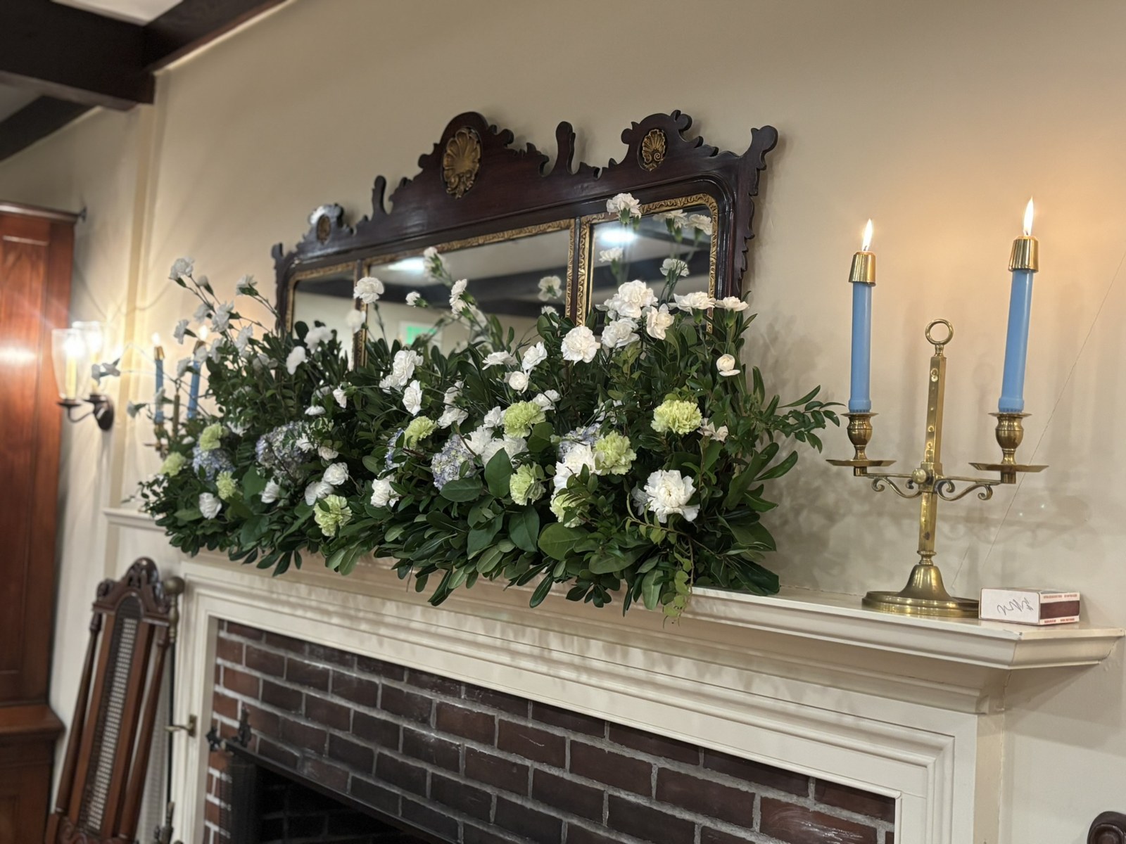 White flowers and greenery across a fireplace mantel beneath an antique mirror