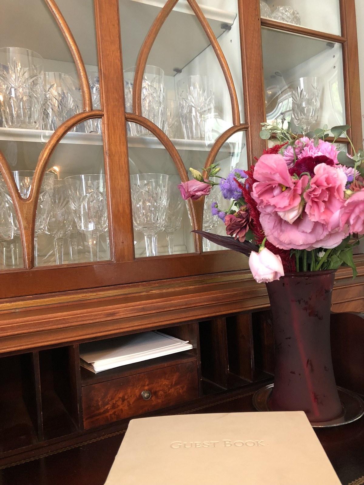 Pink flowers in a tall burgundy vessel beside an antique glass-front cabinet
