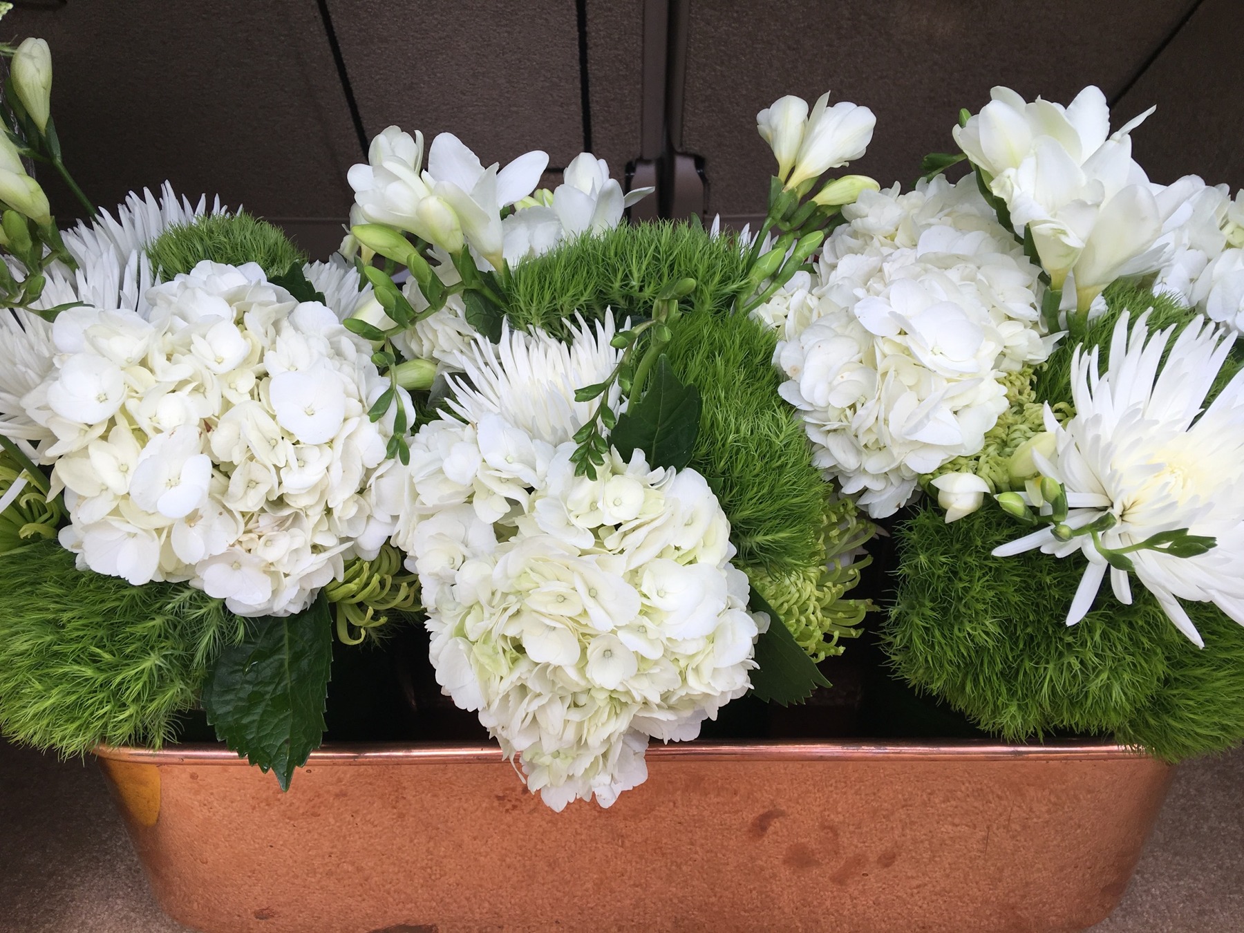 White hydrangeas and other white flowers with green foliage in a long copper vessel