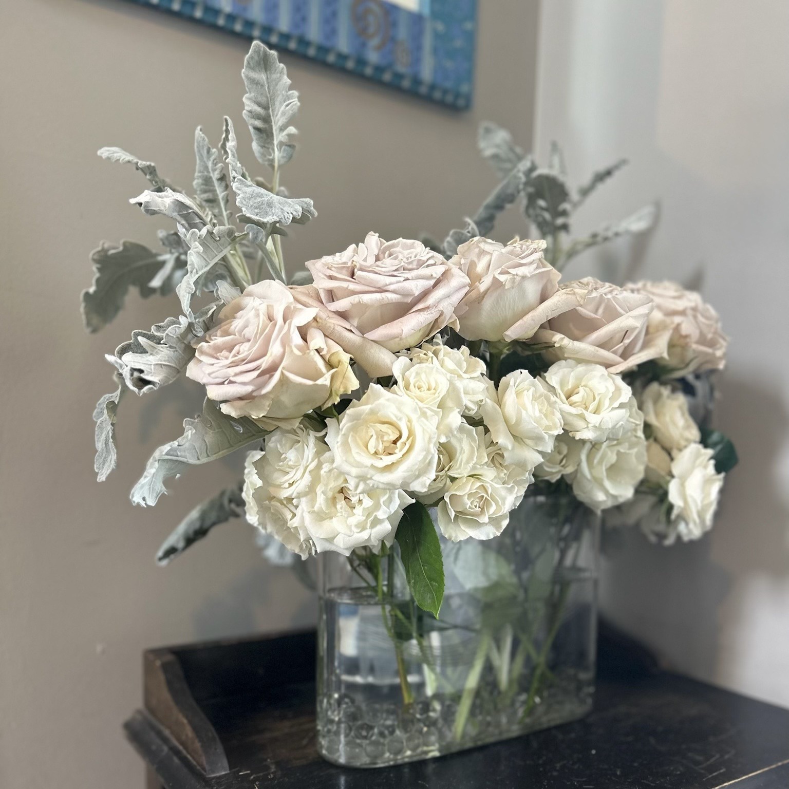 Pale pink and white roses with silvery foliage in a vintage glass vessel on a side table