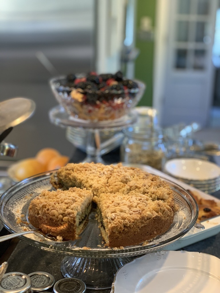 A cake on a raised glass stand beside fruit and breakfast dishes