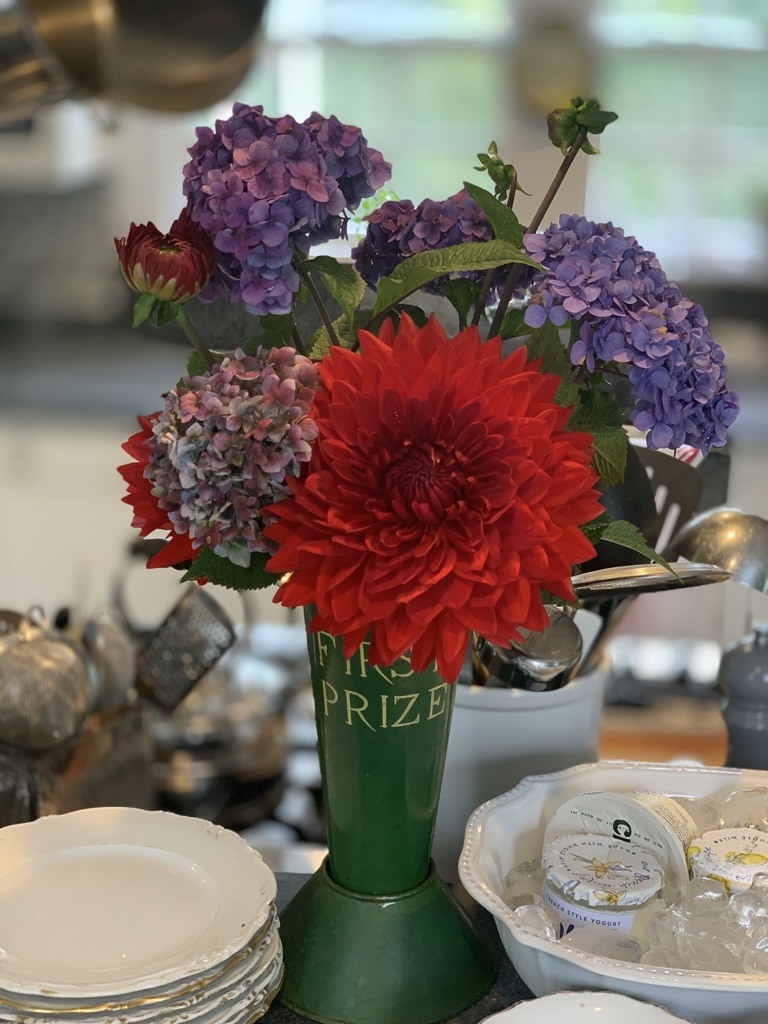 Red and purple flowers in a green trophy-shaped vessel beside stacked plates