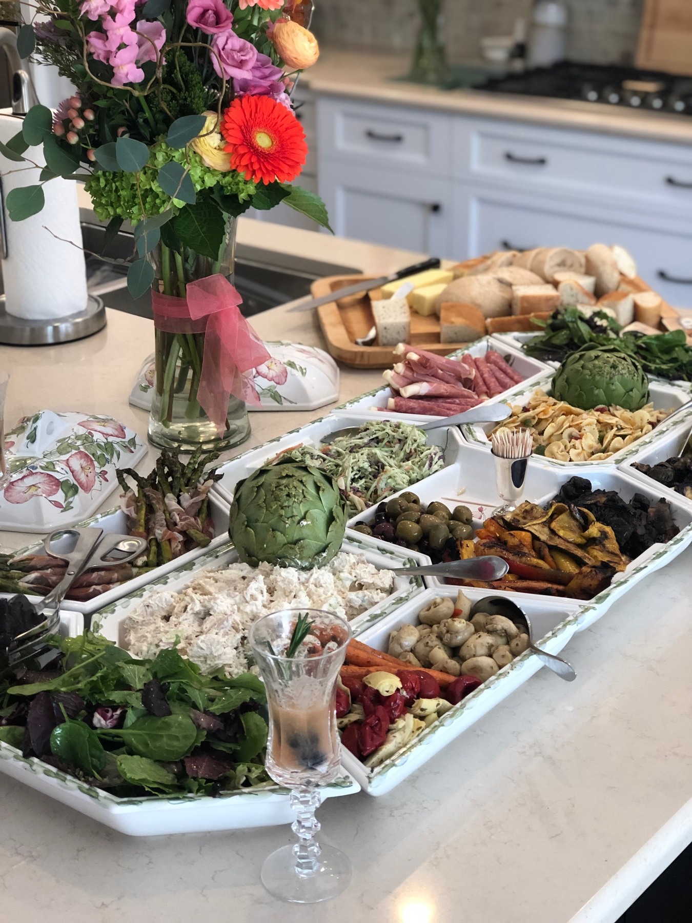 Antipasto platters, salads, vegetables and bread beside colorful flowers on a kitchen island
