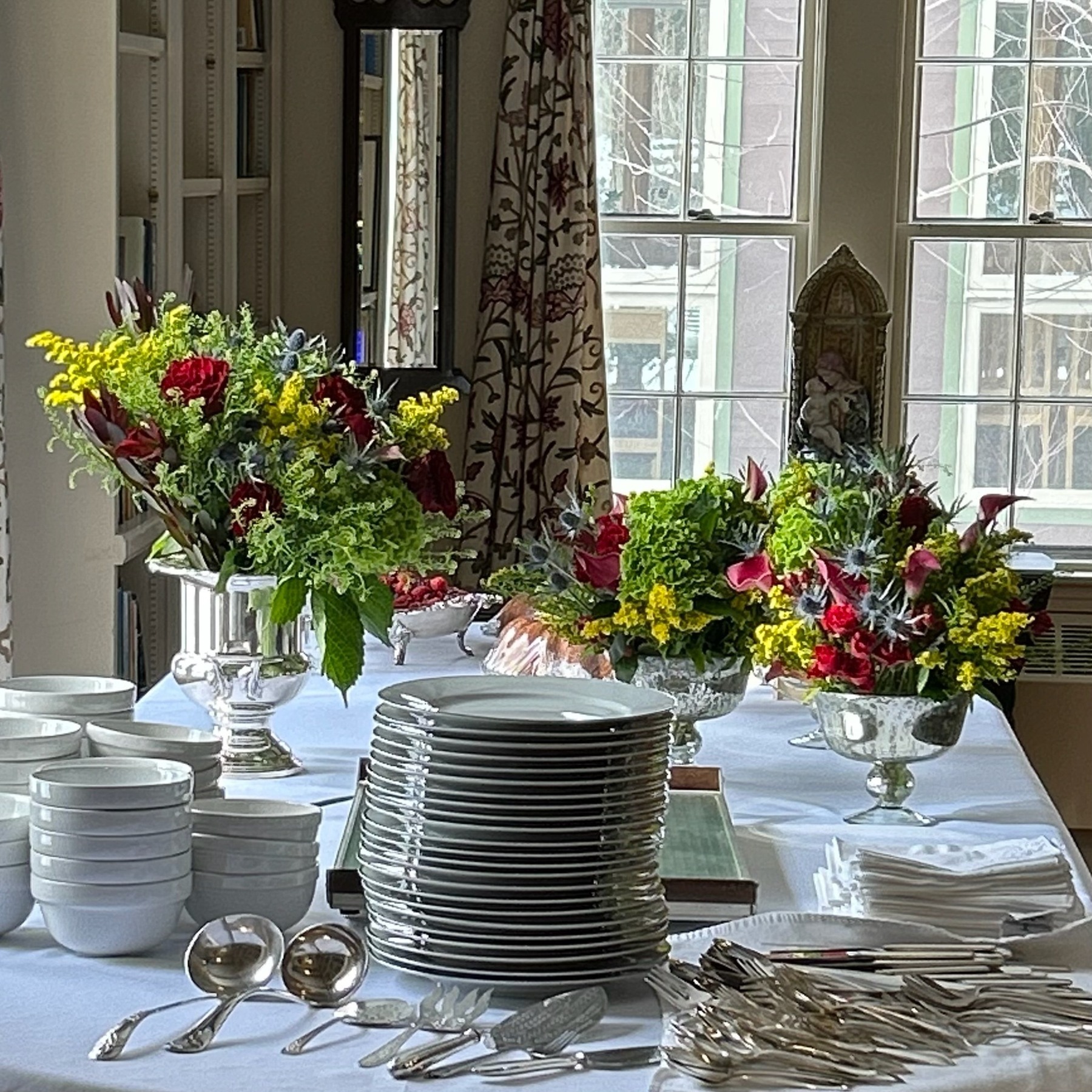 Colorful flowers in silver vessels behind stacks of plates and serving utensils
