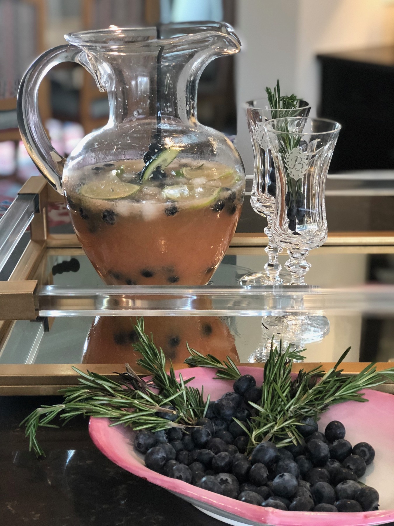 Fruit-filled drink pitcher with crystal glasses, blueberries and rosemary on a mirrored tray