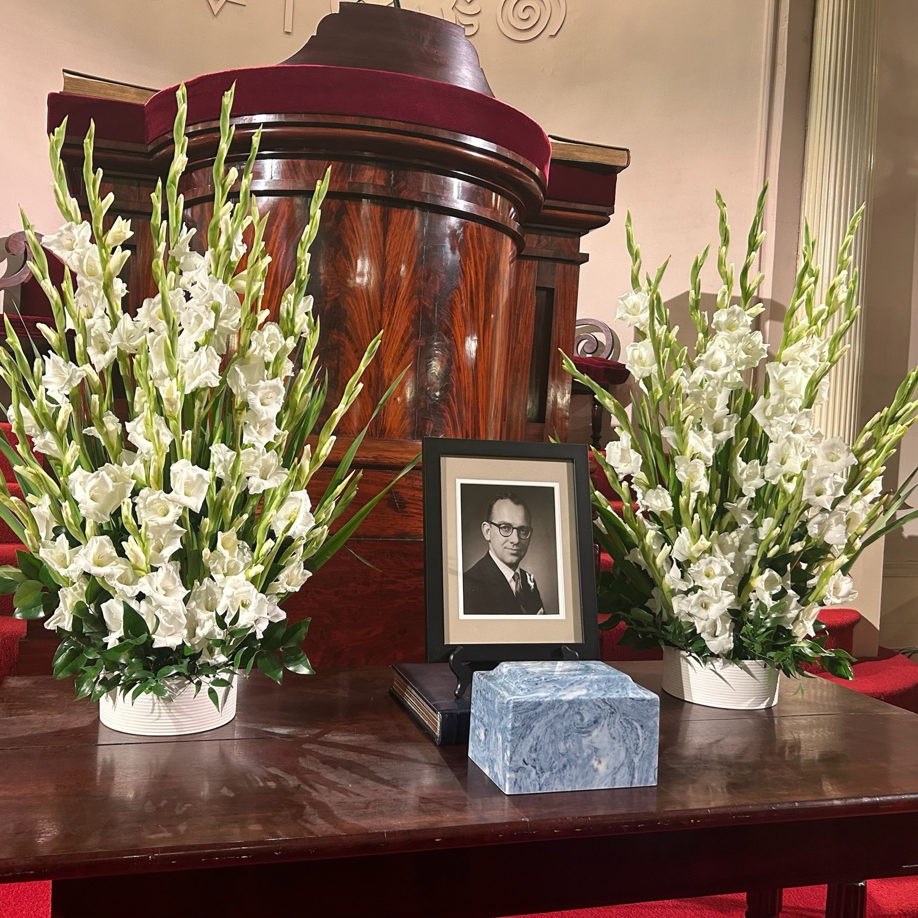 Two arrangements of white gladioli beside a framed portrait at a memorial service