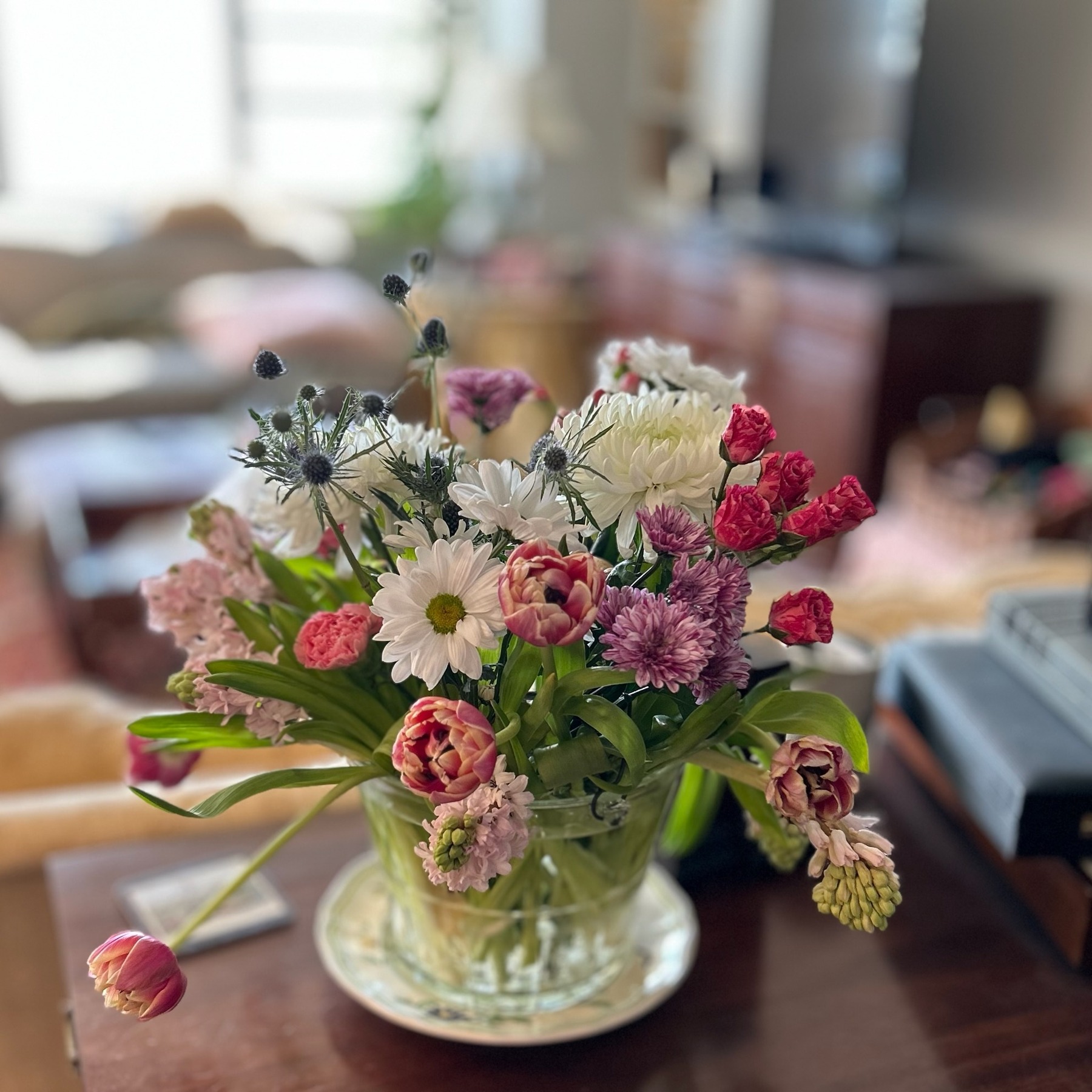 Pink tulips, white daisies, purple flowers and blue thistle in a clear glass vase