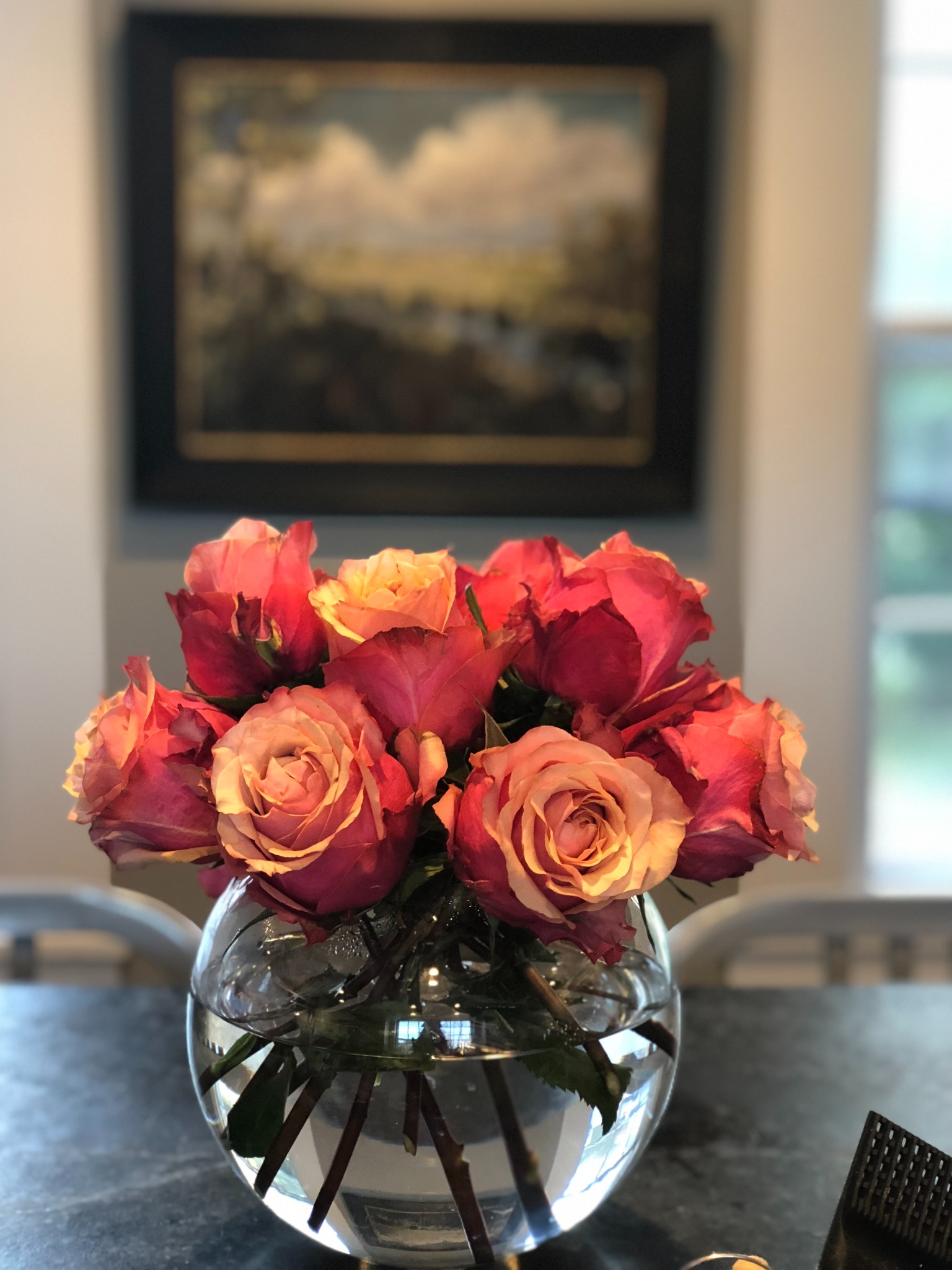 Coral and peach roses in a round glass bowl in front of a framed landscape painting