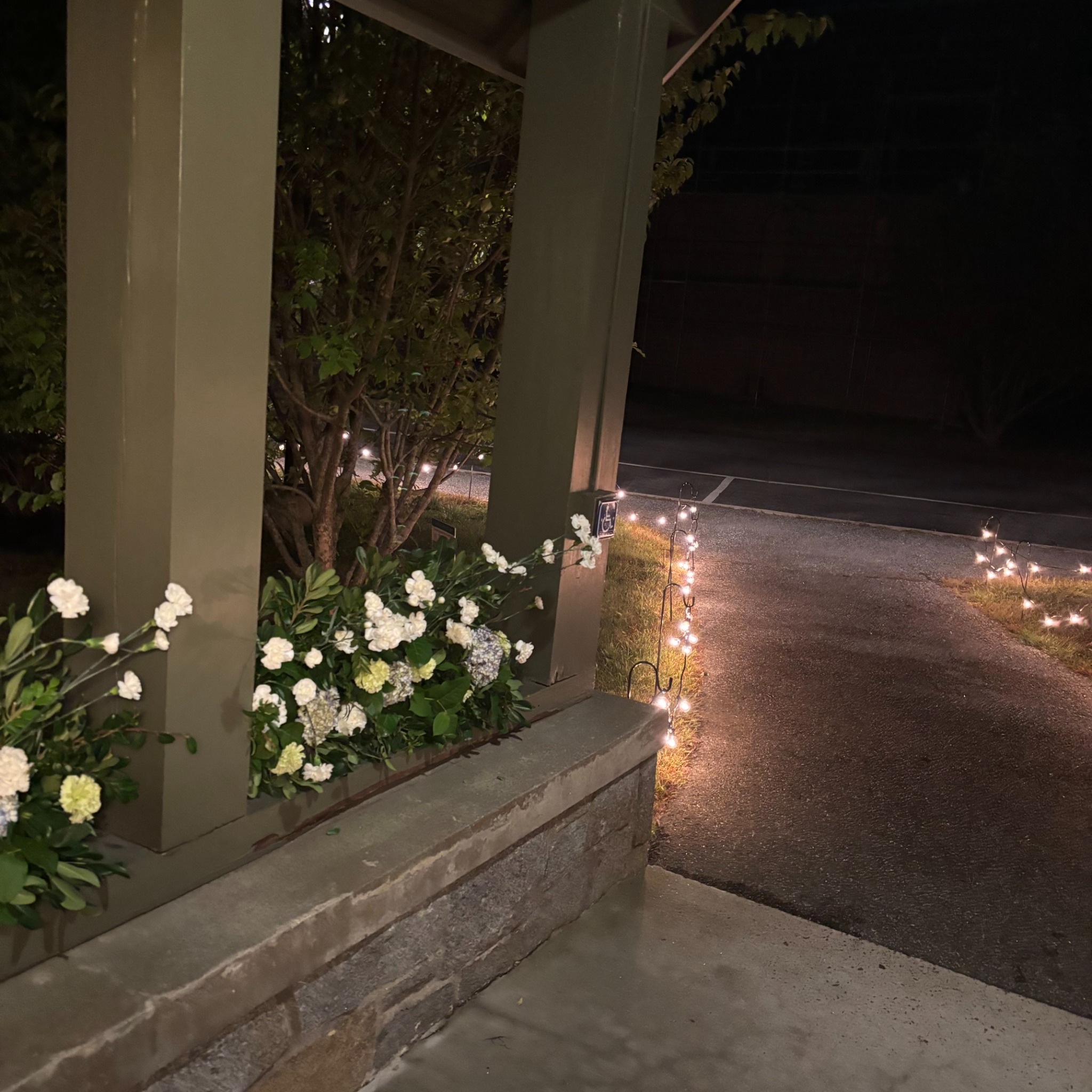 White wedding flowers and warm string lights beside the church entrance and garden path