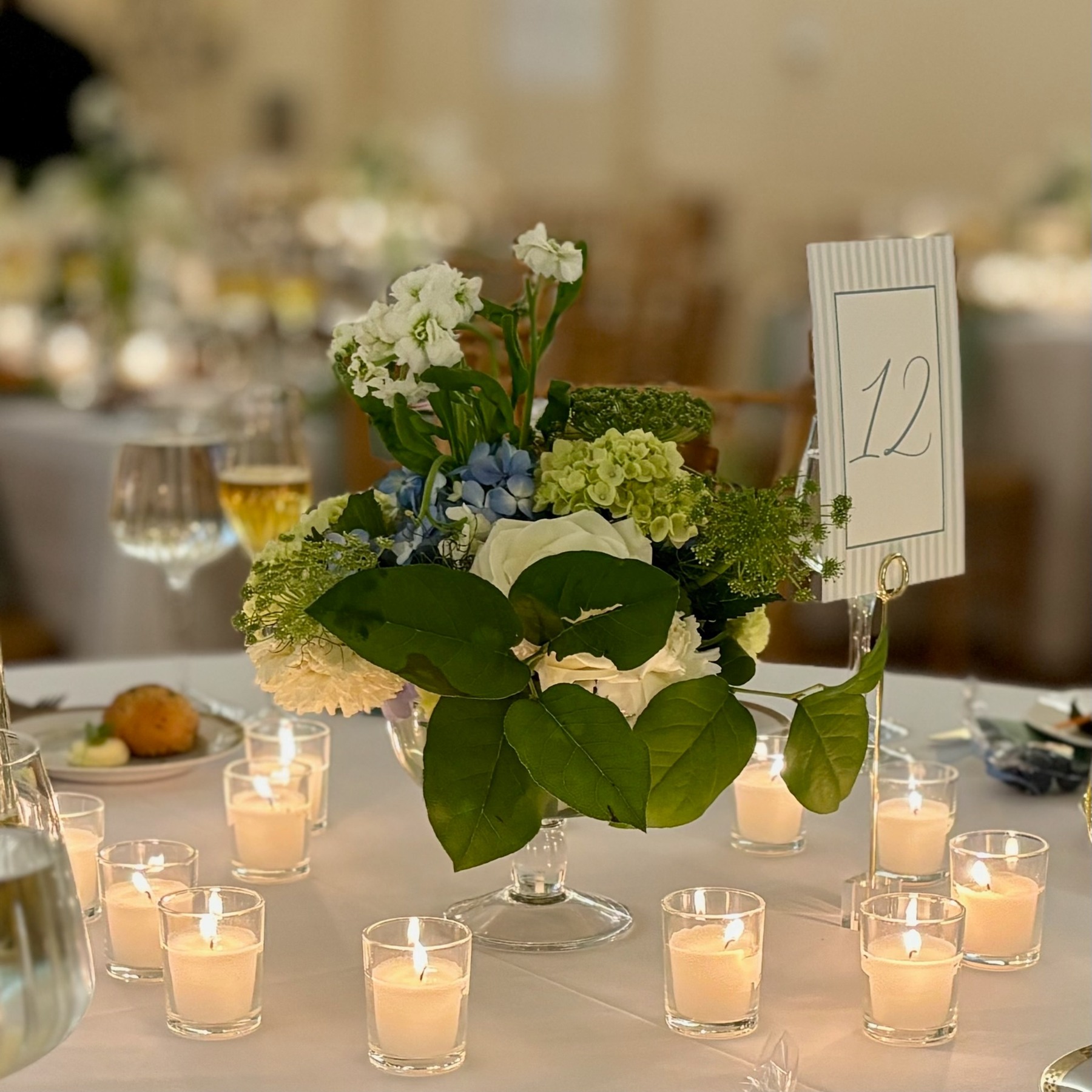 White, blue and green flowers in a footed glass vessel surrounded by candles at a wedding table