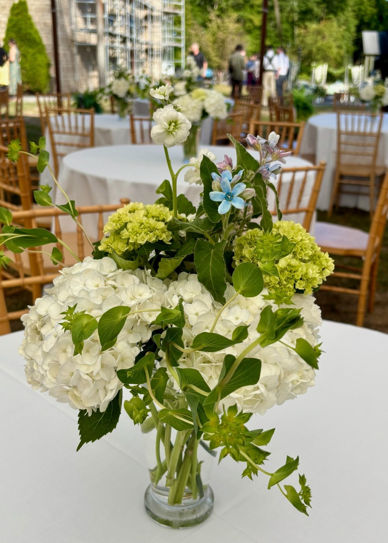 White and green flowers in a glass vessel with garden reception tables behind