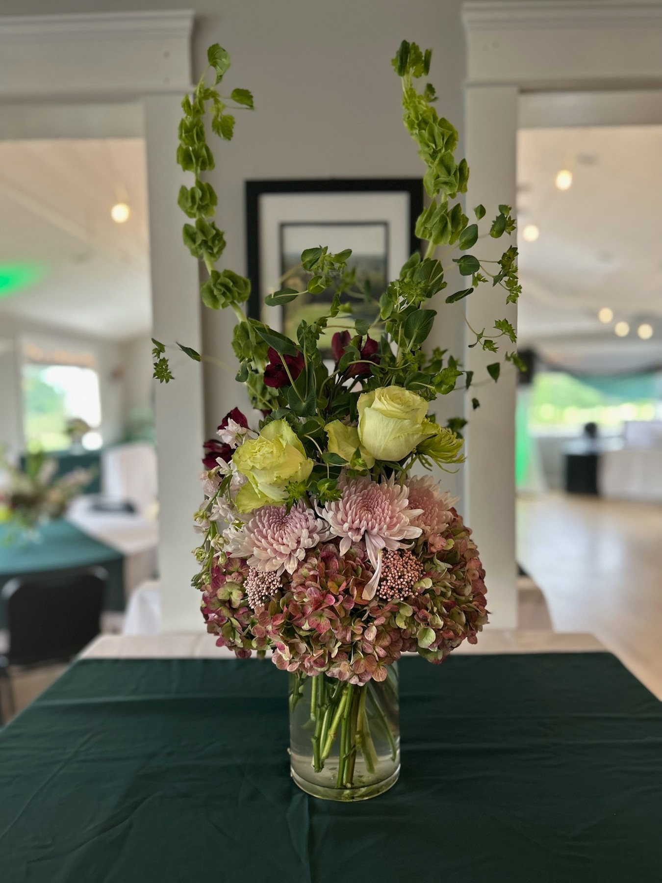 Tall green stems, pink hydrangeas and pale roses in a glass vessel on a covered table
