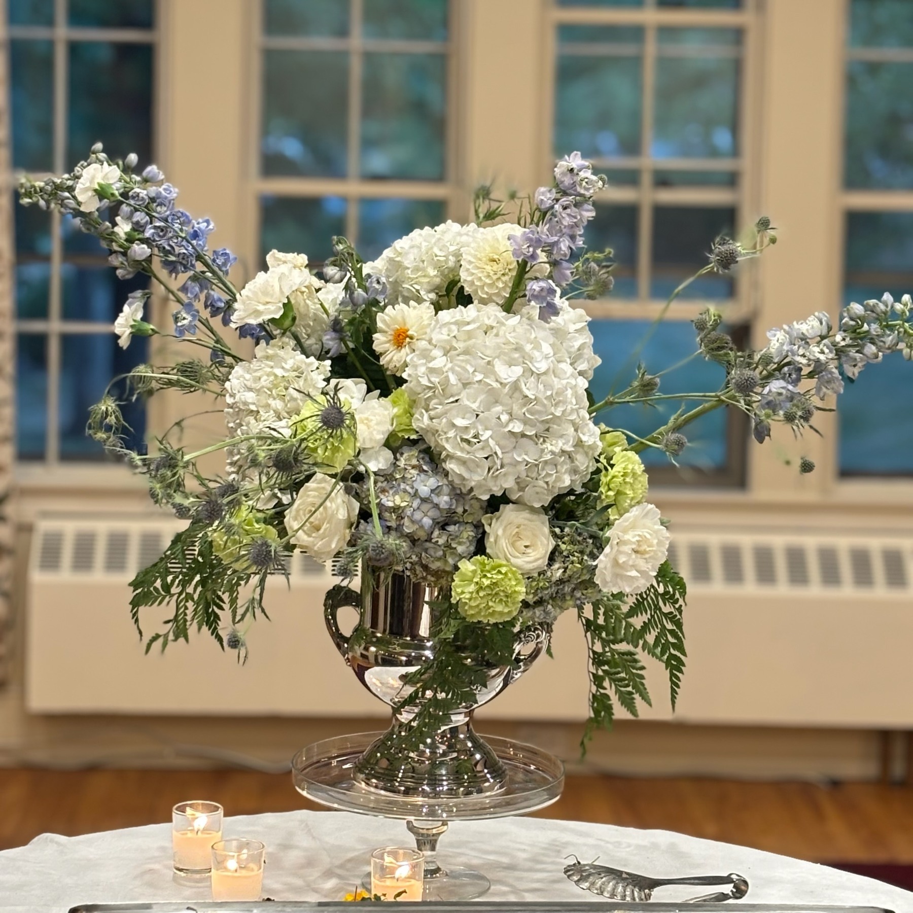 White and pale blue flowers with trailing greenery in a footed silver vessel